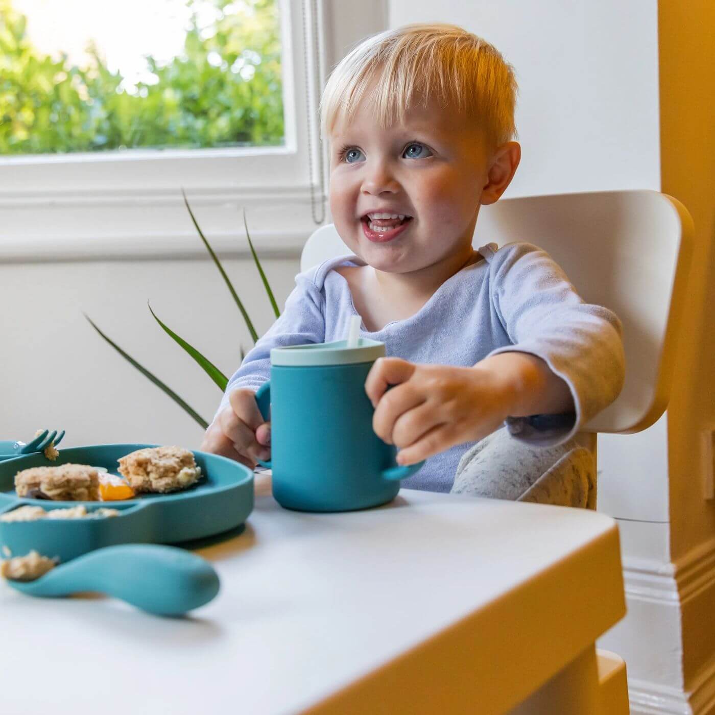 A smiling toddler holds a blue TUM TUM silicone sippy cup with dual handles and a straw lid, designed for easy gripping and independent drinking. The toddler sits at a table with matching blue dinnerware, enjoying a meal in a comfortable high chair.