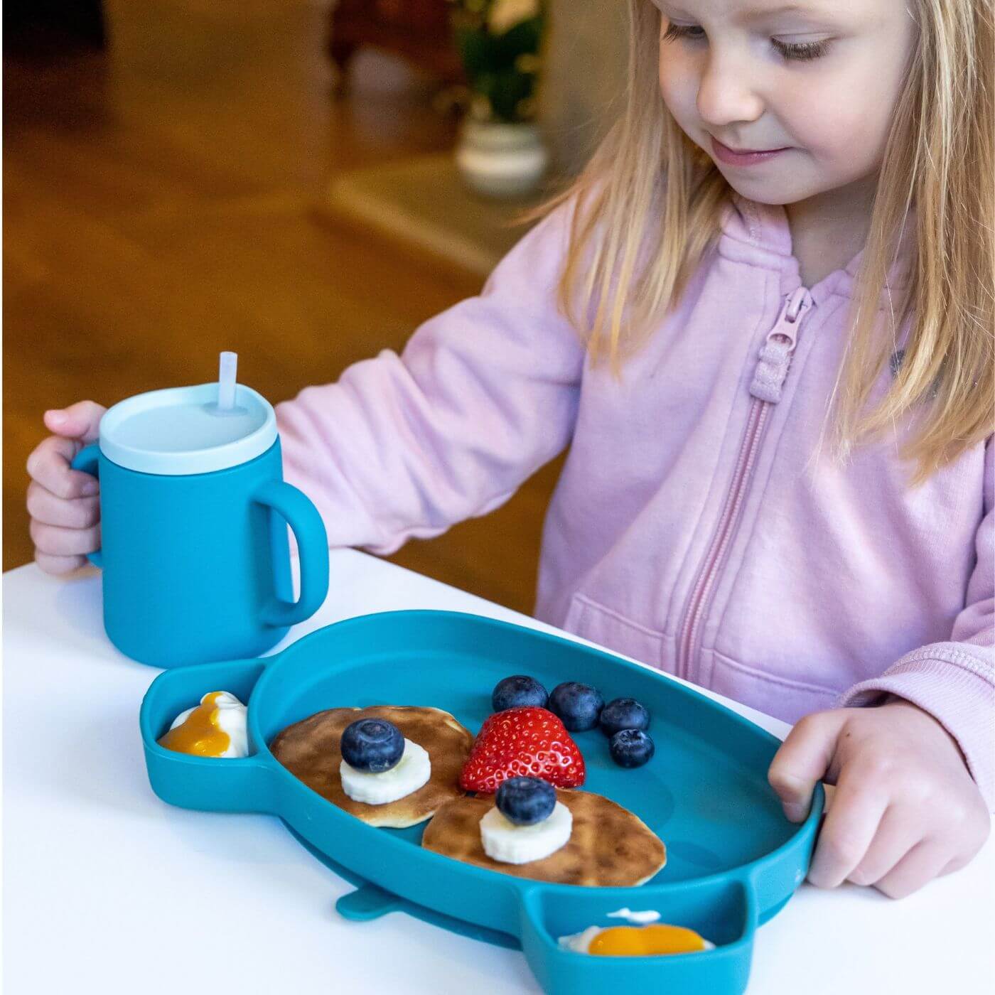 A child holds a blue TUM TUM silicone sippy cup with a straw lid, alongside a matching divided plate filled with a healthy meal of pancakes, fruits, and sauces. The cup’s dual handles offer easy grip, making it perfect for toddlers and young children learning to drink independently.