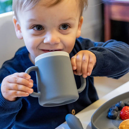 A toddler is drinking from a grey TUM TUM silicone sippy cup with dual handles, designed for easy gripping. 