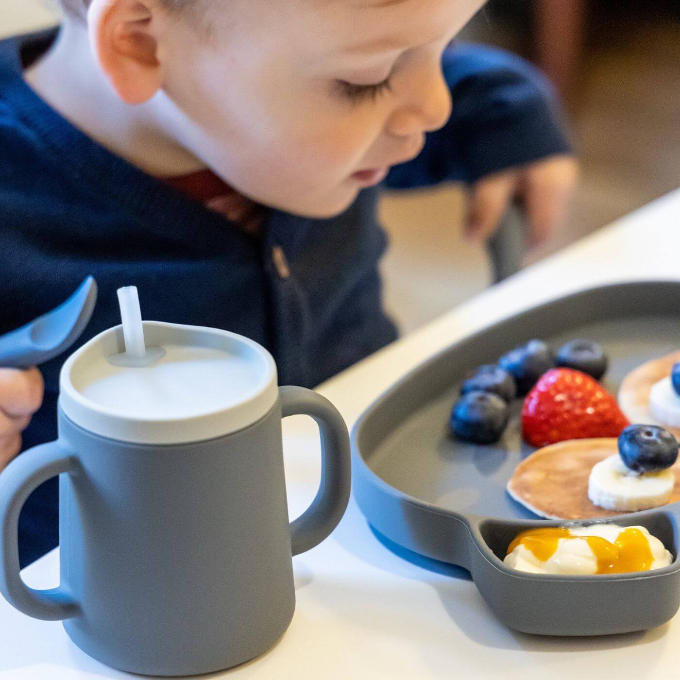 A toddler enjoys a meal featuring pancakes and fruit, using a grey TUM TUM silicone sippy cup with dual handles and a straw lid. The child is holding a matching silicone spoon, and the plate is neatly divided for different foods, perfect for independent eating and drinking.