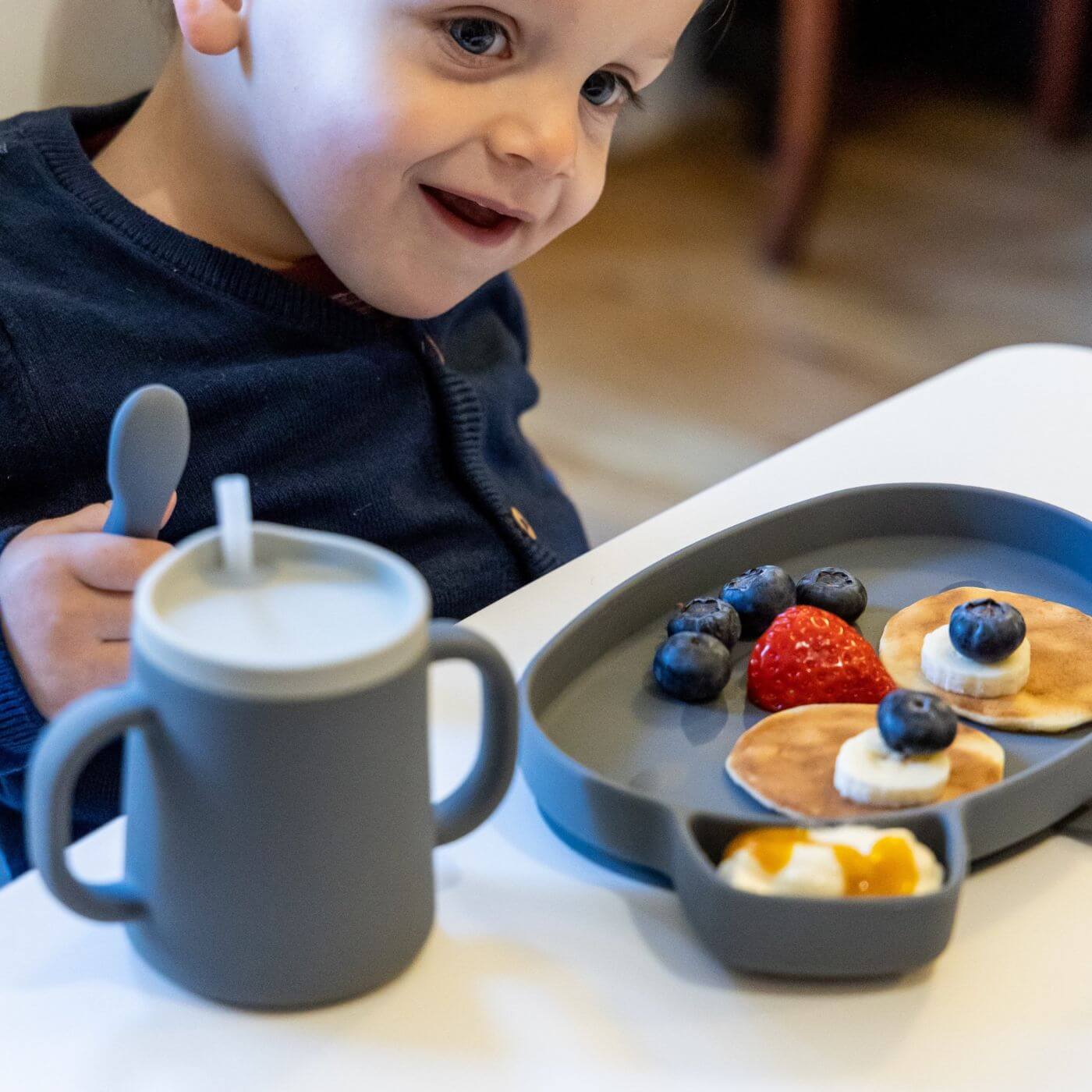 A toddler is enjoying a meal of pancakes and fruit while using a grey TUM TUM silicone sippy cup with dual handles and a straw lid. The child holds a matching silicone spoon, with the plate thoughtfully divided for easy self-feeding, promoting independent eating and drinking.