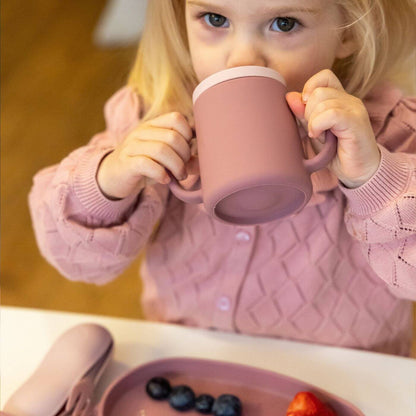 A  toddler sips a drink from a pink TUM TUM silicone sippy cup with dual handles and a straw lid.