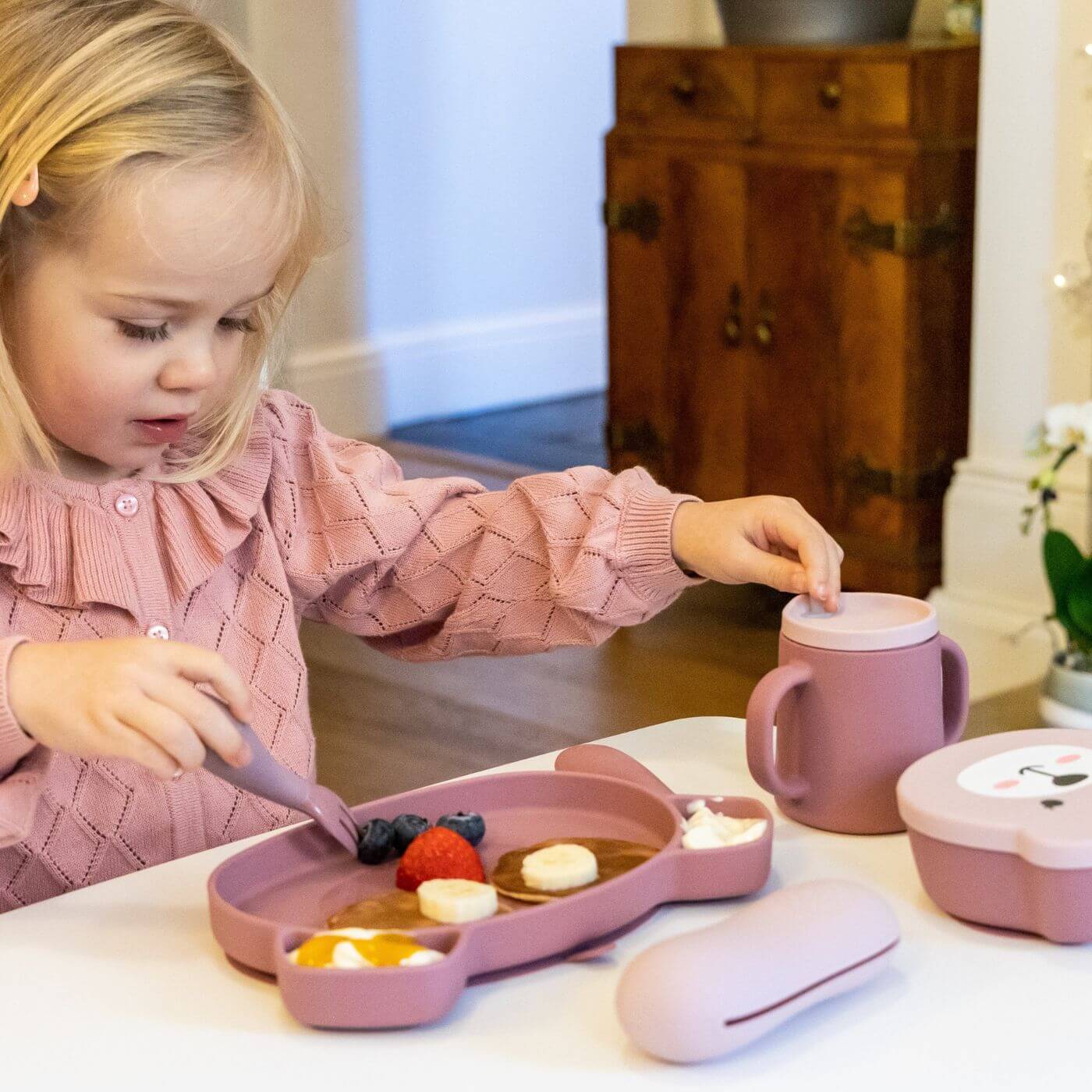 A toddler is enjoying a meal of pancakes and fruit while using a pink TUM TUM silicone sippy cup with dual handles and a straw lid. The child holds a matching silicone spoon, with the plate thoughtfully divided for easy self-feeding, promoting independent eating and drinking.