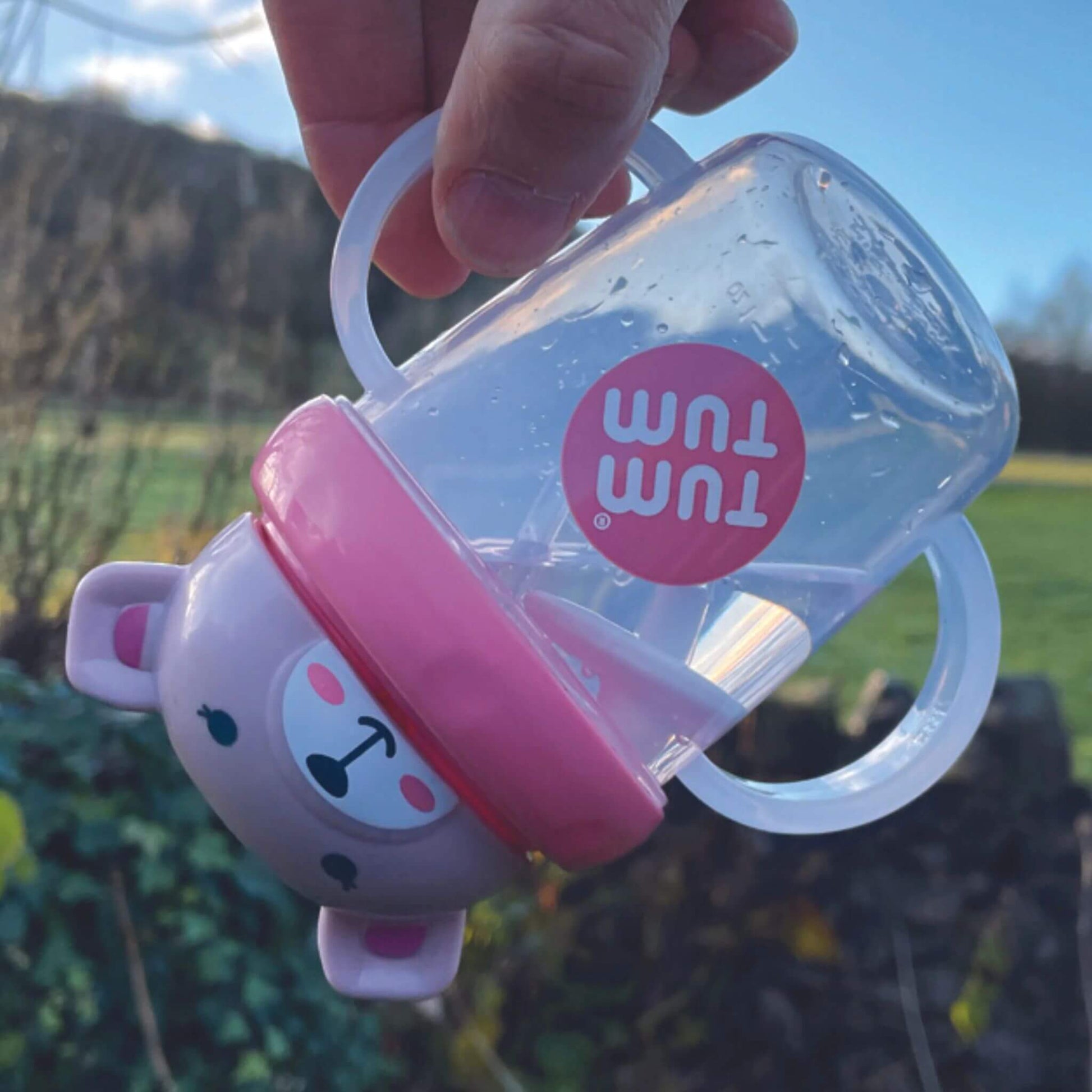 A TUM TUM Tippy Up Cup featuring a Betsy bear lid with two clear handles, shown being tipped upside down. The weighted straw inside follows the liquid, making it easy for toddlers to drink from any angle. The BPA-free design ensures safety and spill-free drinking while on the go.