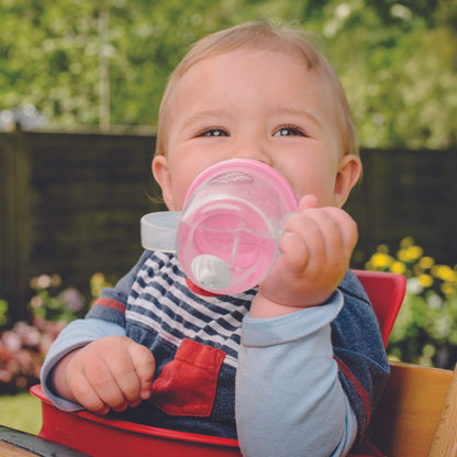 A baby happily drinking from a TUM TUM Tippy Up Cup with a pink bear lid. The cup has clear handles and a weighted straw that follows the liquid, allowing the baby to drink comfortably from any angle. Ideal for toddlers learning to drink independently.