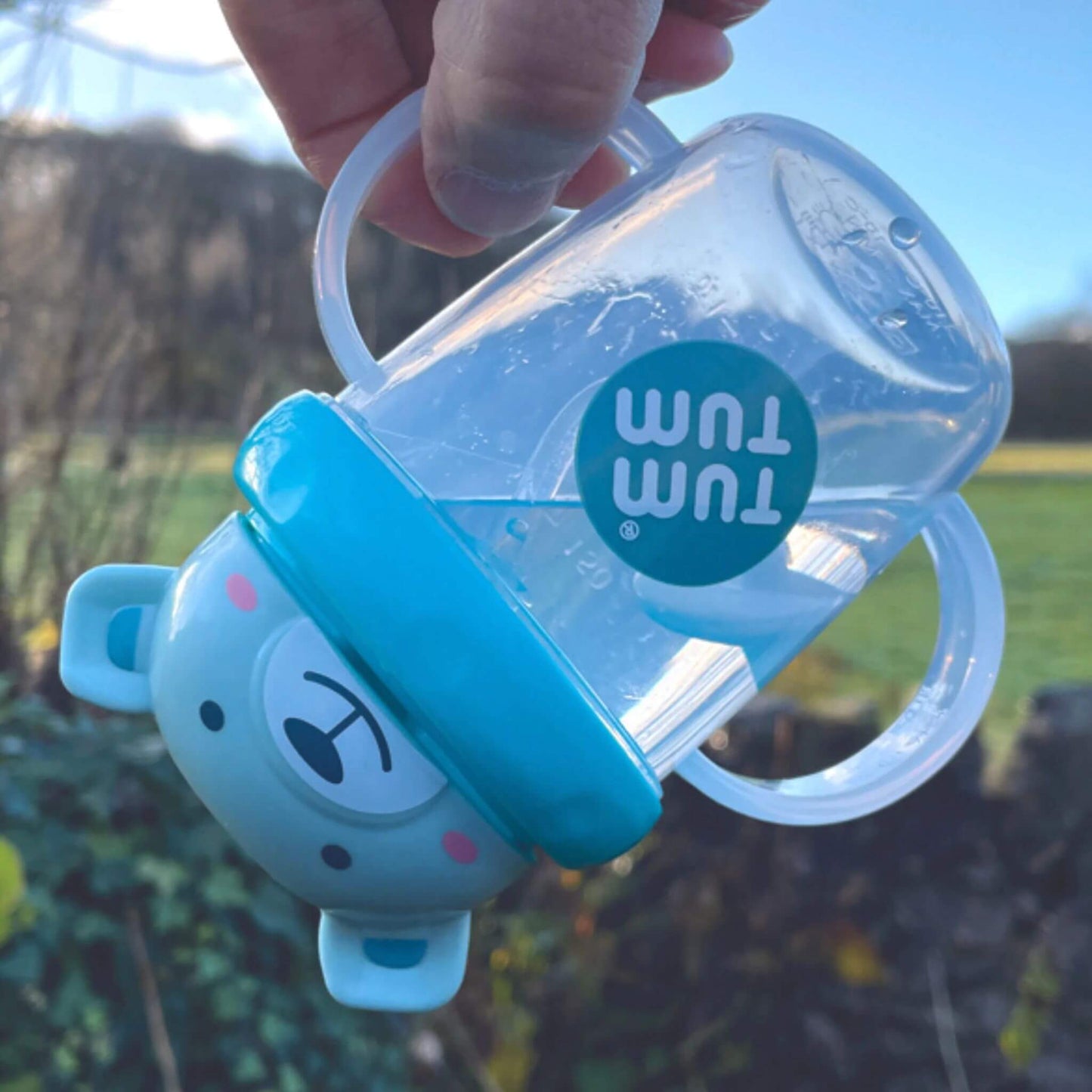 A TUM TUM Tippy Up Cup featuring a Boris bear lid with two clear handles, shown being tipped upside down. The weighted straw inside follows the liquid, making it easy for toddlers to drink from any angle. The BPA-free design ensures safety and spill-free drinking while on the go.