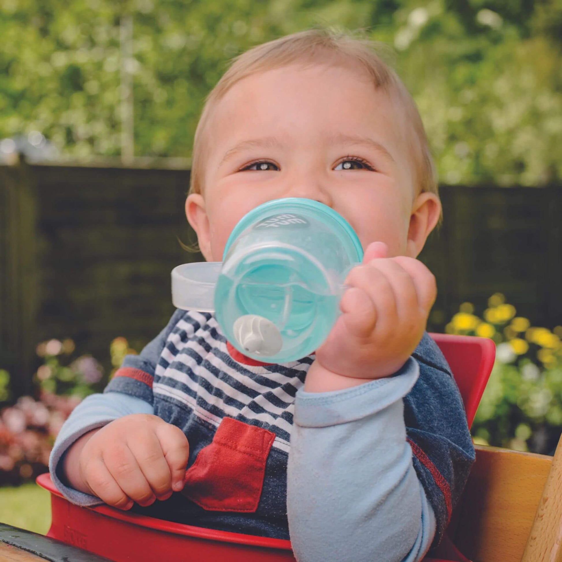 A baby happily drinking from a TUM TUM Tippy Up Cup with a blue bear lid. The cup has clear handles and a weighted straw that follows the liquid, allowing the baby to drink comfortably from any angle. Ideal for toddlers learning to drink independently.