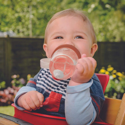 A baby happily drinking from a TUM TUM Tippy Up Cup. The cup has clear handles and a weighted straw that follows the liquid, allowing the baby to drink comfortably from any angle. Ideal for toddlers learning to drink independently.