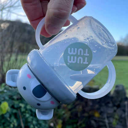 A TUM TUM Tippy Up Cup featuring a Kev Koala lid with two clear handles, shown being tipped upside down. The weighted straw inside follows the liquid, making it easy for toddlers to drink from any angle. The BPA-free design ensures safety and spill-free drinking while on the go.