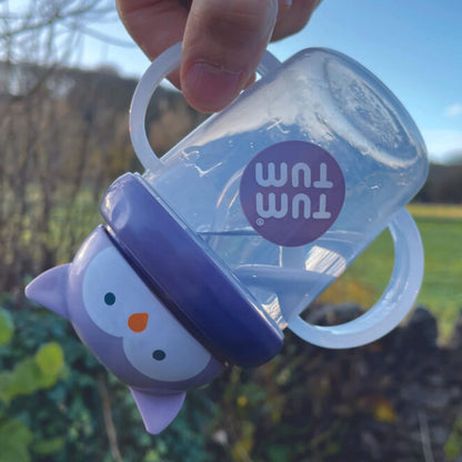 A TUM TUM Tippy Up Cup featuring a cute owl lid with two clear handles, shown being tipped upside down. The weighted straw inside follows the liquid, making it easy for toddlers to drink from any angle. The BPA-free design ensures safety and spill-free drinking while on the go.