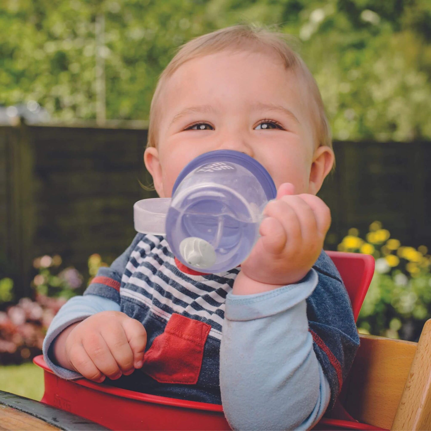 A baby happily drinking from a TUM TUM Tippy Up Cup. The cup has clear handles and a weighted straw that follows the liquid, allowing the baby to drink comfortably from any angle. Ideal for toddlers learning to drink independently.