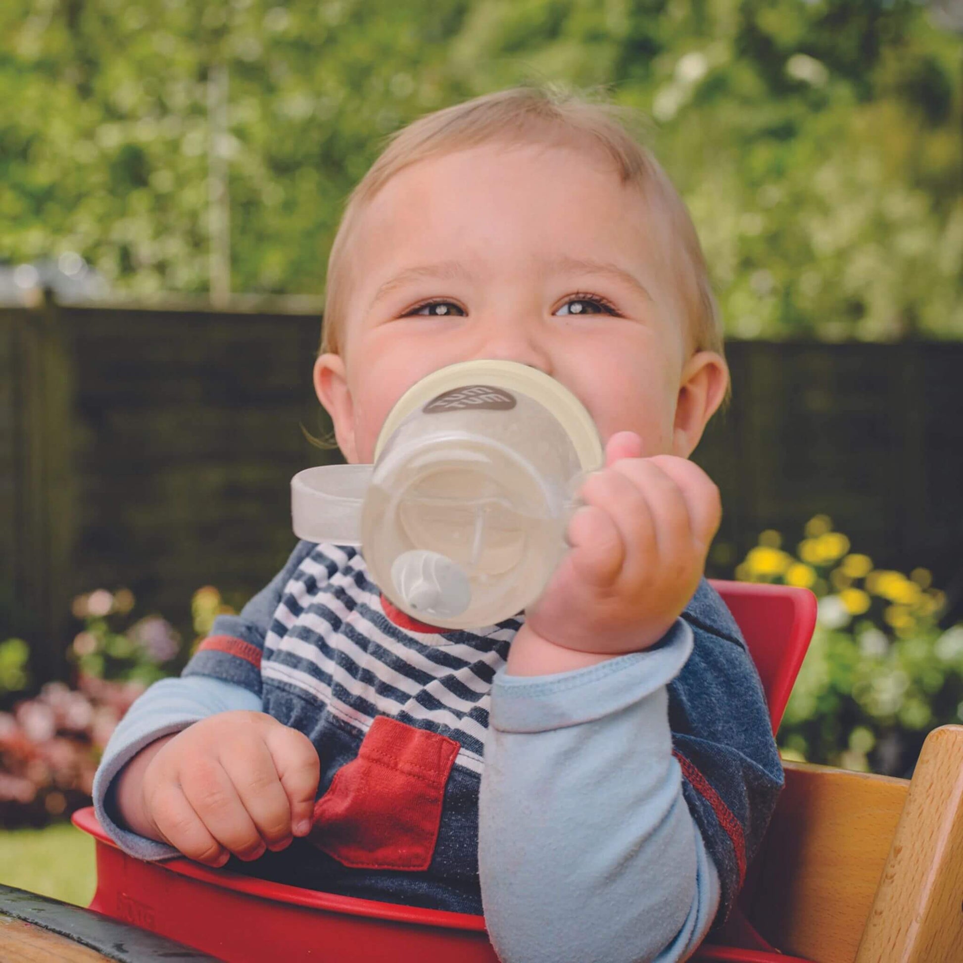 A baby happily drinking from a TUM TUM Tippy Up Cup. The cup has clear handles and a weighted straw that follows the liquid, allowing the baby to drink comfortably from any angle. Ideal for toddlers learning to drink independently.