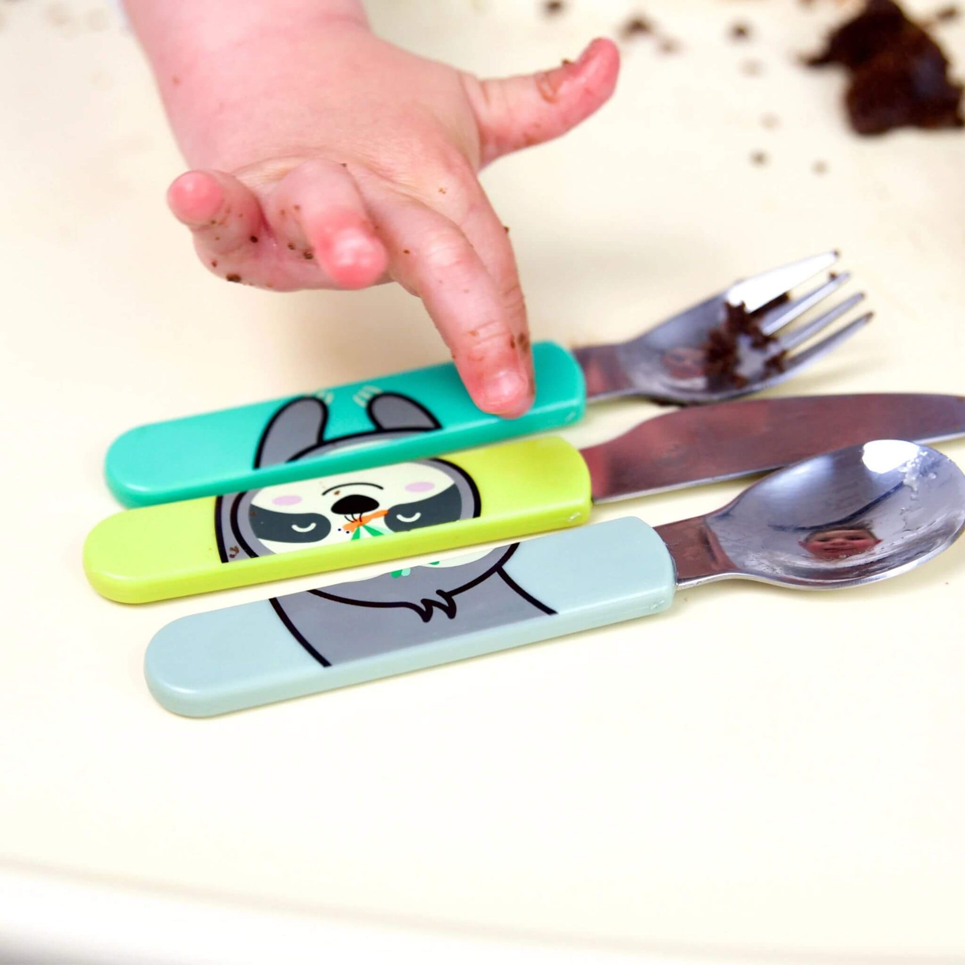 A child using the TUM TUM Easy Scoop Cutlery Set, featuring Stanley Sloth handles on the spoon, knife, and fork. Designed for easy grip and self-feeding, with a fun and colourful design.