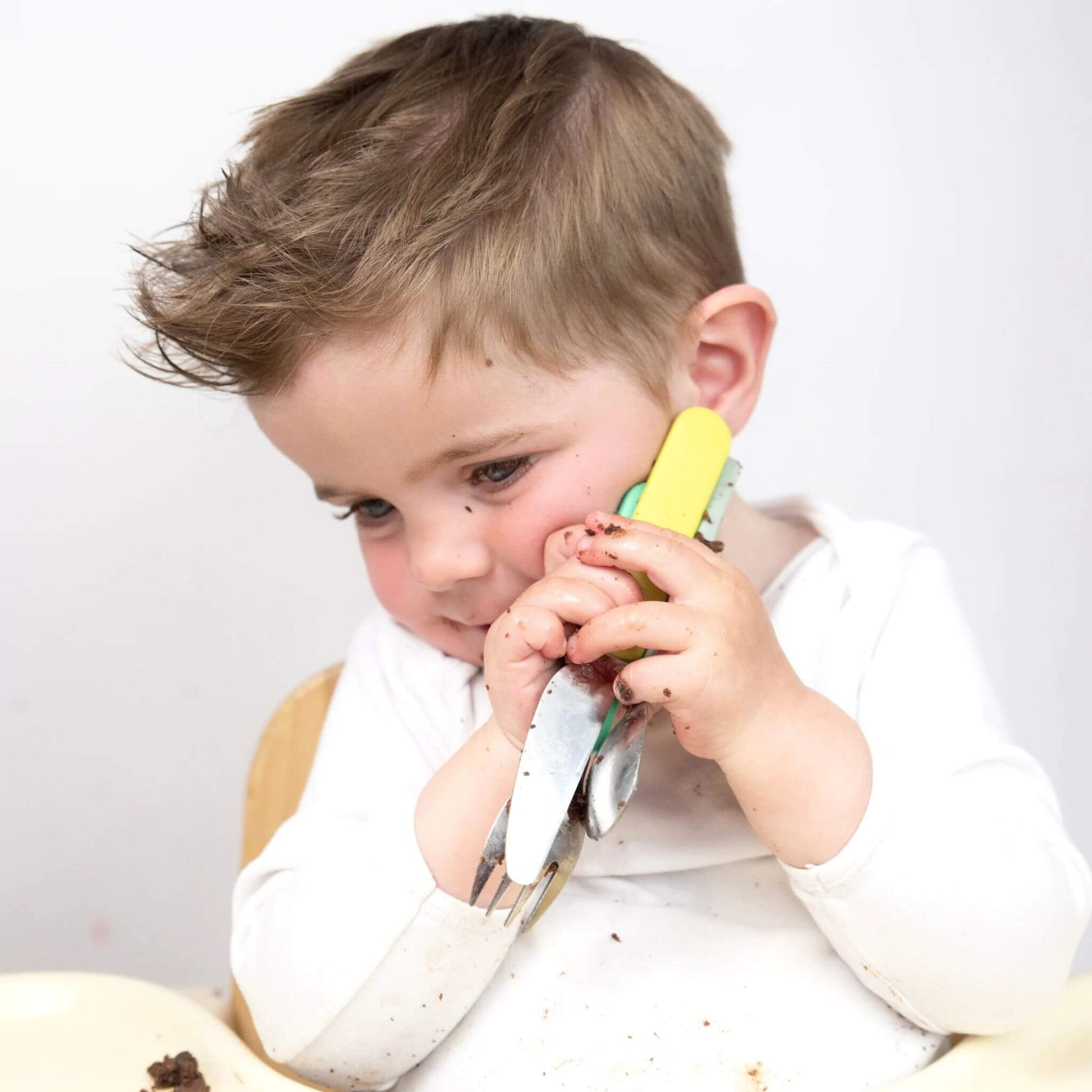 A toddler holding the TUM TUM Easy Scoop Cutlery Set, featuring Stanley Sloth handles on the fork and spoon. The set is designed for easy grip and self-feeding.