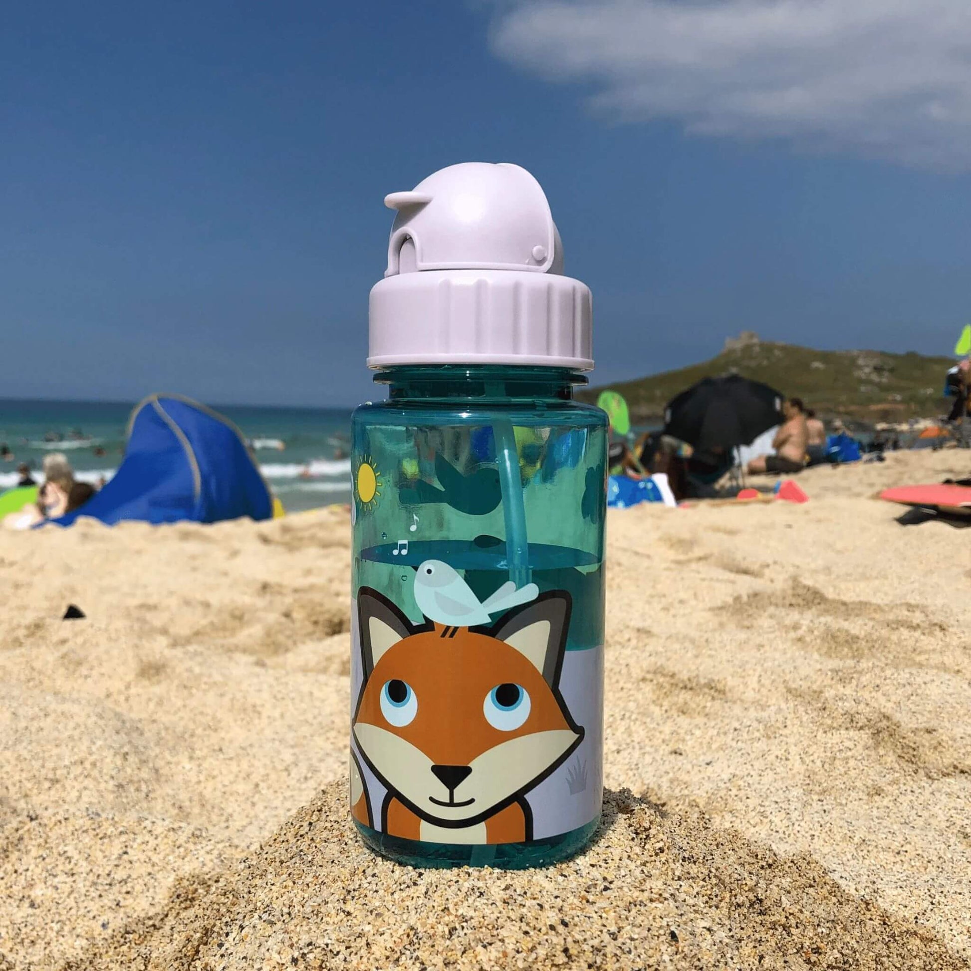 TUM TUM water bottle with Felicity Fox design, featuring a flip top lid and built-in straw, placed on a sandy beach with people in the background.