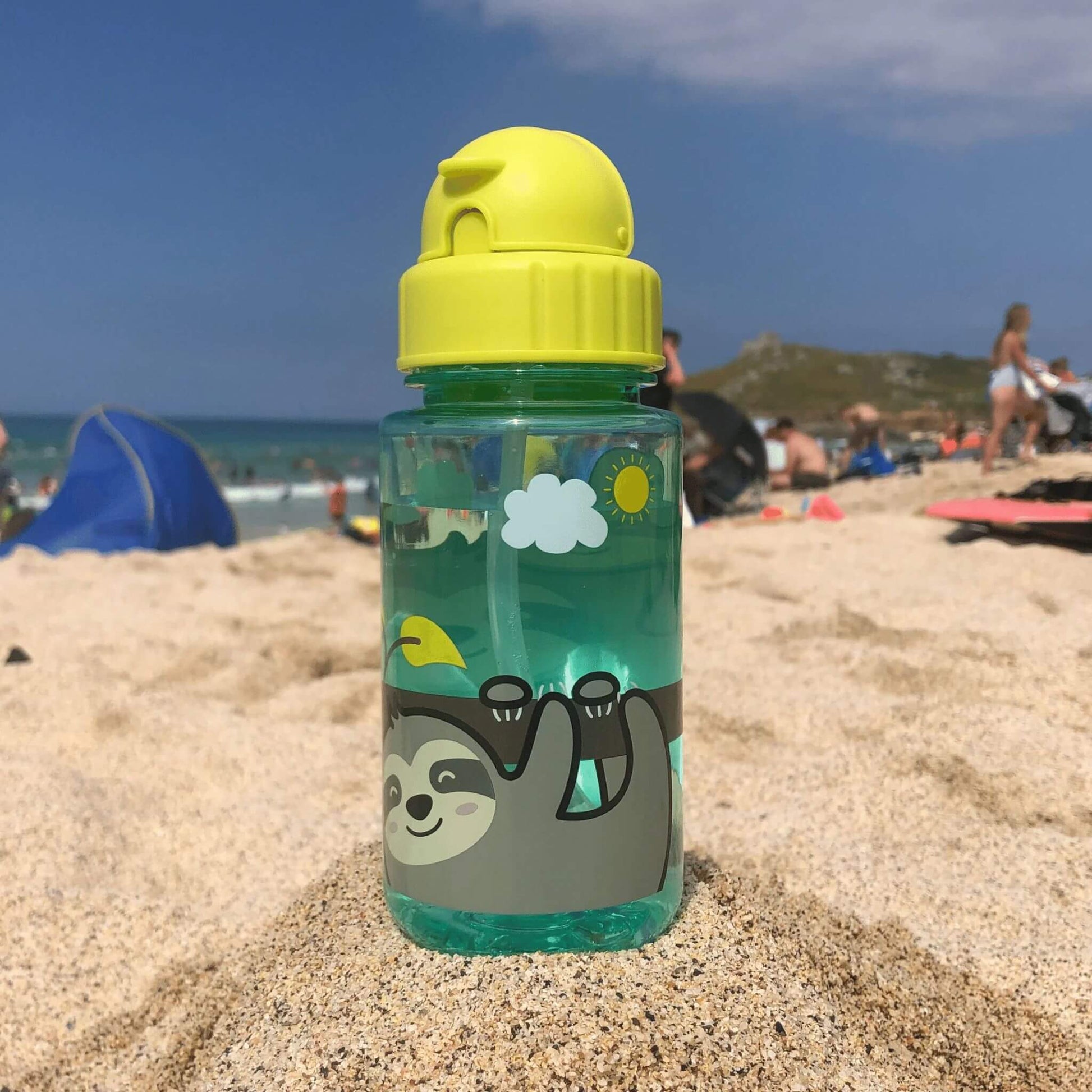 TUM TUM water bottle with sloth design, featuring a flip top lid and built-in straw, placed on a sandy beach with people in the background.