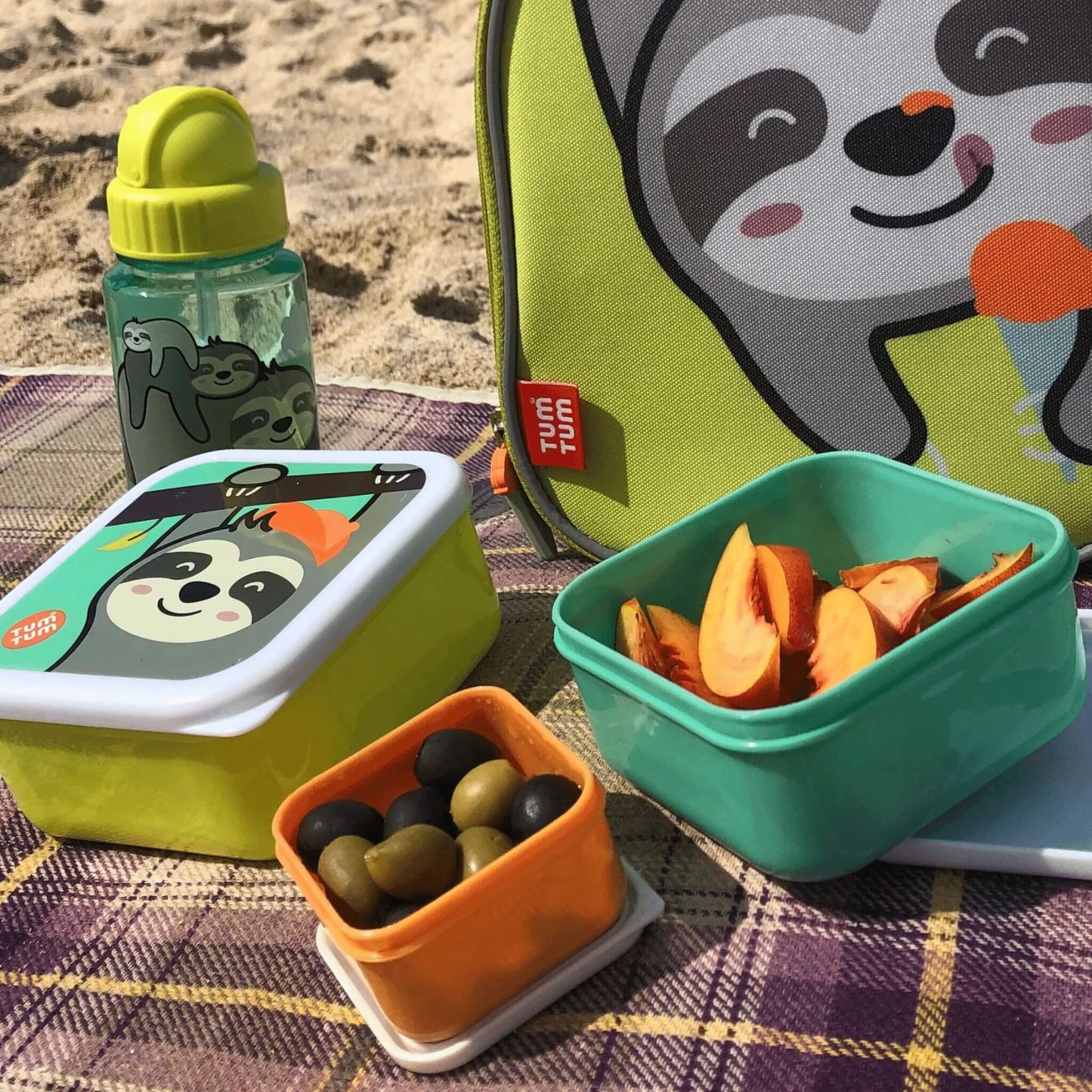 TUM TUM water bottle with Stanley sloth design, alongside matching snack boxes and lunch bag, placed on a blanket at the beach, perfect for kids' meals.