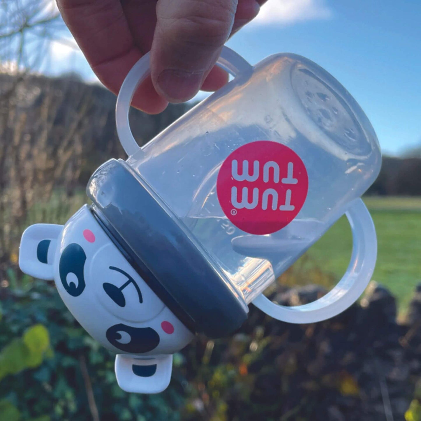 A TUM TUM Tippy Up Cup featuring a panda bear lid with two clear handles, shown being tipped upside down. The weighted straw inside follows the liquid, making it easy for toddlers to drink from any angle. The BPA-free design ensures safety and spill-free drinking while on the go.