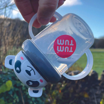 A TUM TUM Tippy Up Cup featuring a panda bear lid with two clear handles, shown being tipped upside down. The weighted straw inside follows the liquid, making it easy for toddlers to drink from any angle. The BPA-free design ensures safety and spill-free drinking while on the go.