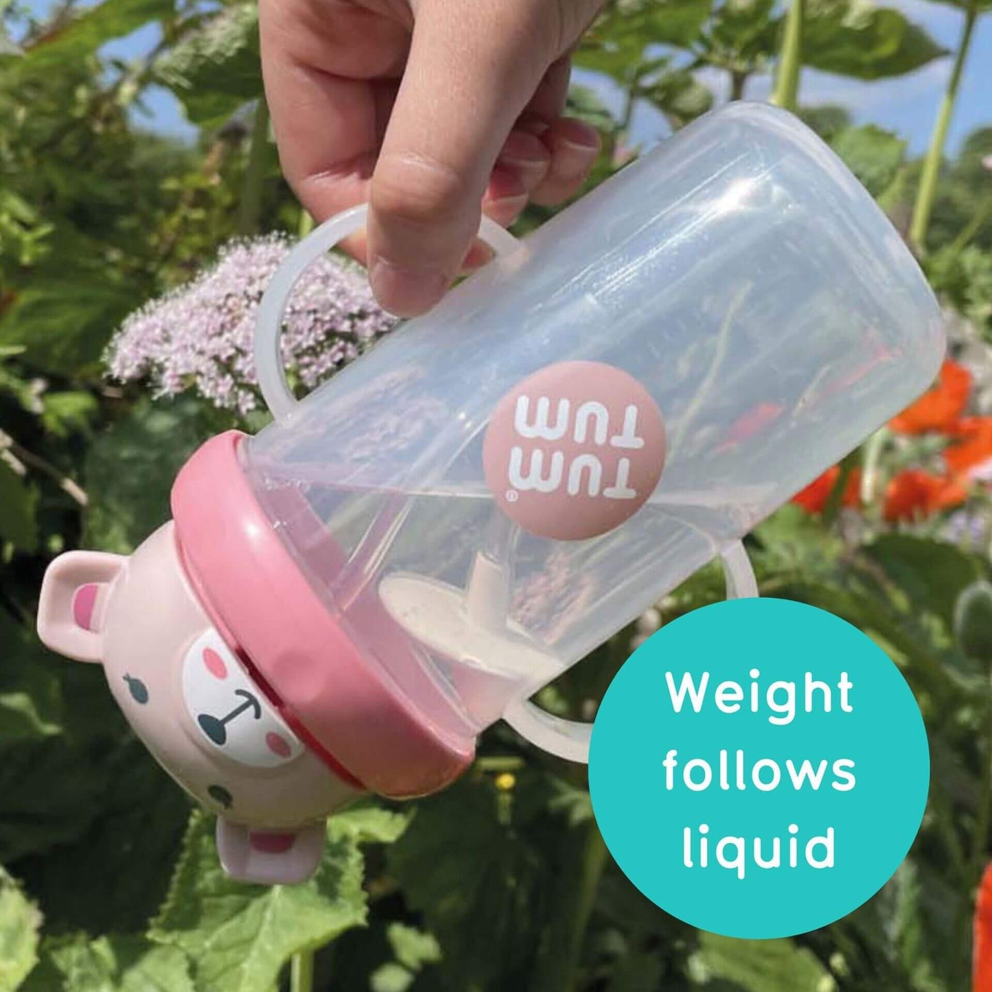 A hand holding a clear TUM TUM sippy cup with a pink bear-shaped lid, tilted to show the weighted straw following the liquid inside. The cup is BPA-free with dual handles, designed for easy drinking from any angle.