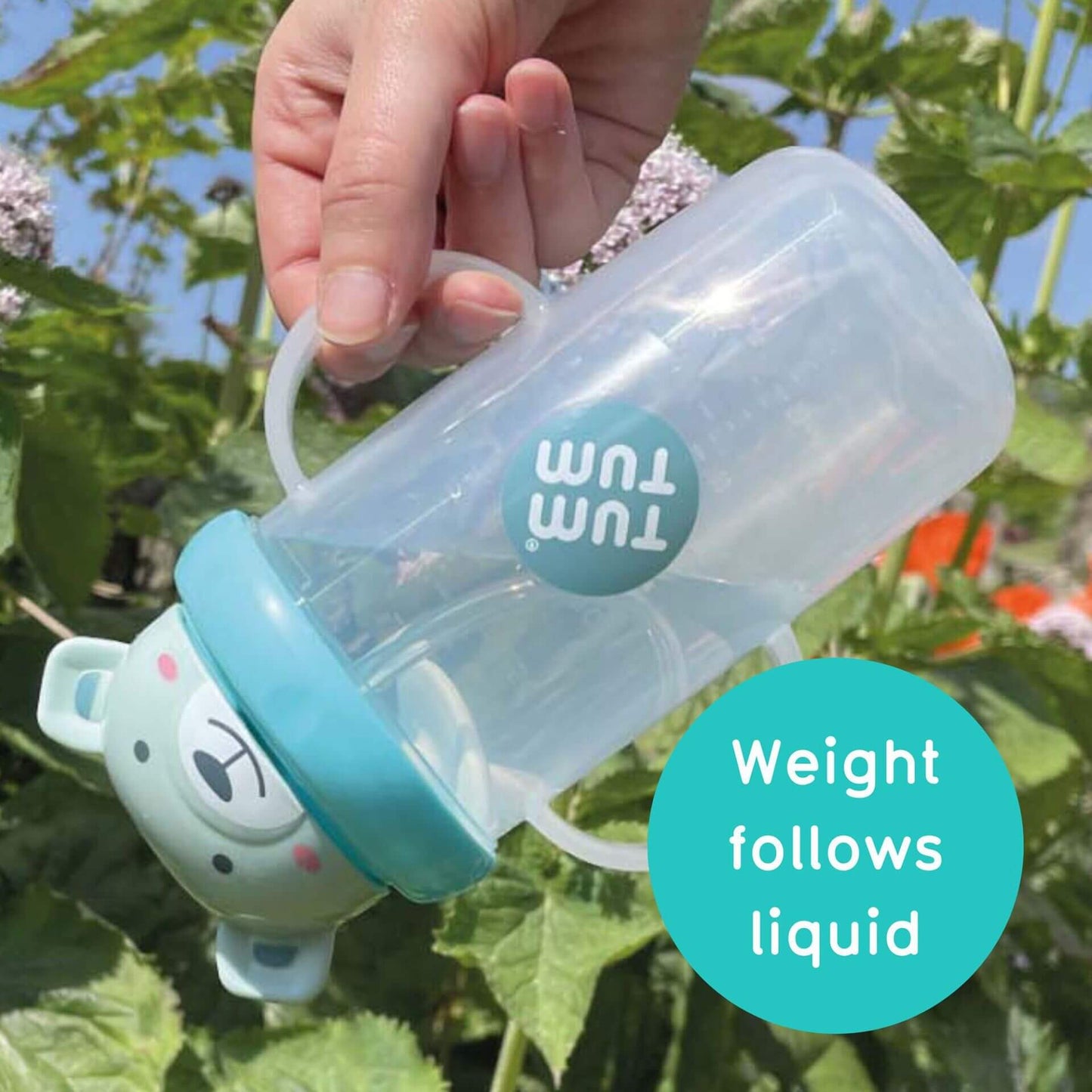 A hand holding a clear TUM TUM sippy cup with a blue bear-shaped lid, tilted to show the weighted straw following the liquid inside. The cup is BPA-free with dual handles, designed for easy drinking from any angle.