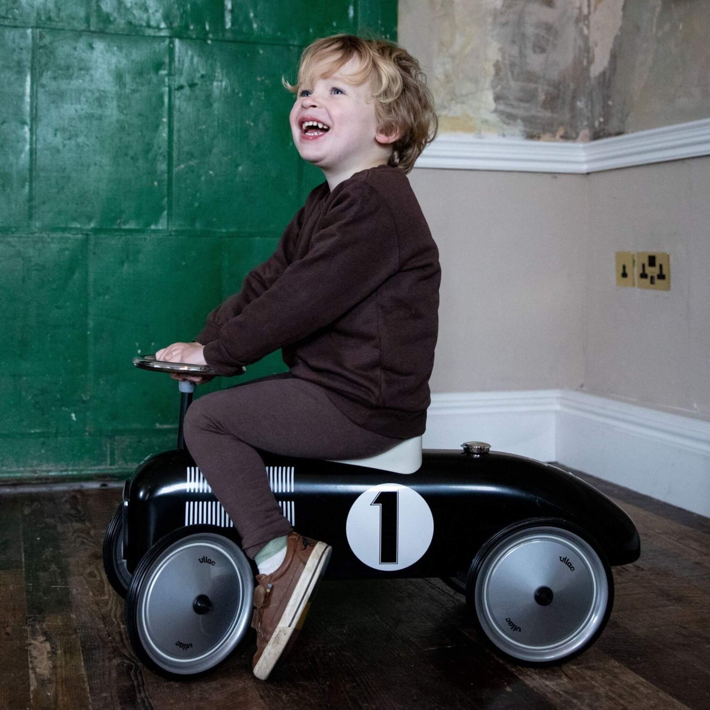 Child riding the Vilac Classic Metal Car in black with a white seat and number 1 detail, smiling joyfully against a rustic green backdrop.