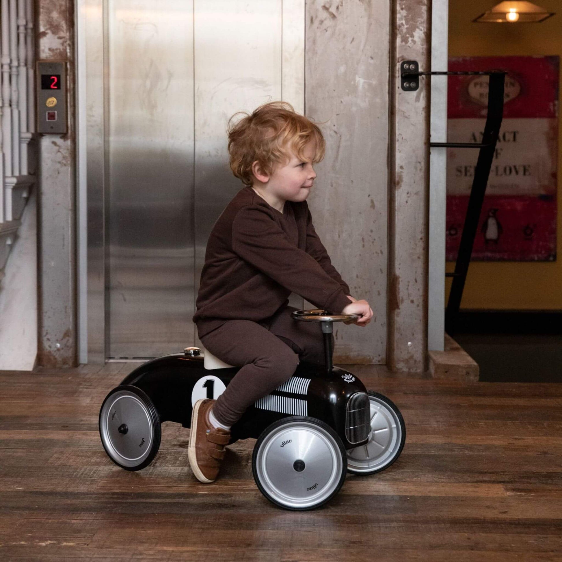 Child riding the Vilac Classic Metal Car in black with a white seat, number 1 detail, and silver steering wheel, set against an industrial-style background.