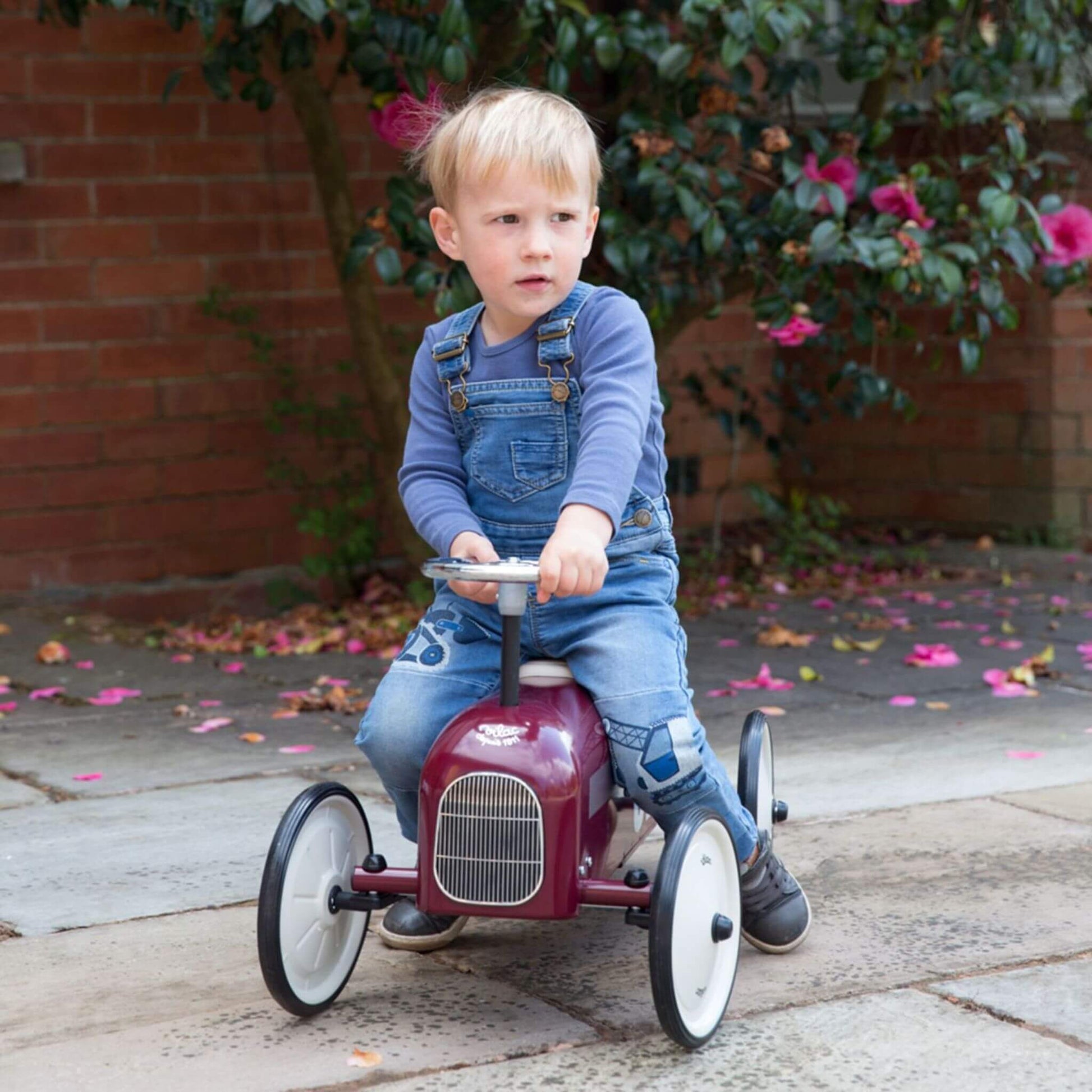 Child riding the Vilac Classic Metal Car in burgundy with a white seat and silver steering wheel, outdoors surrounded by blooming flowers.