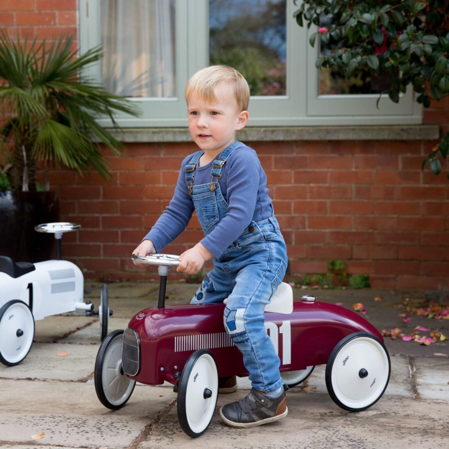 Child riding the Vilac Classic Metal Car in burgundy with white seat and silver steering wheel, outdoors near a brick house and greenery.