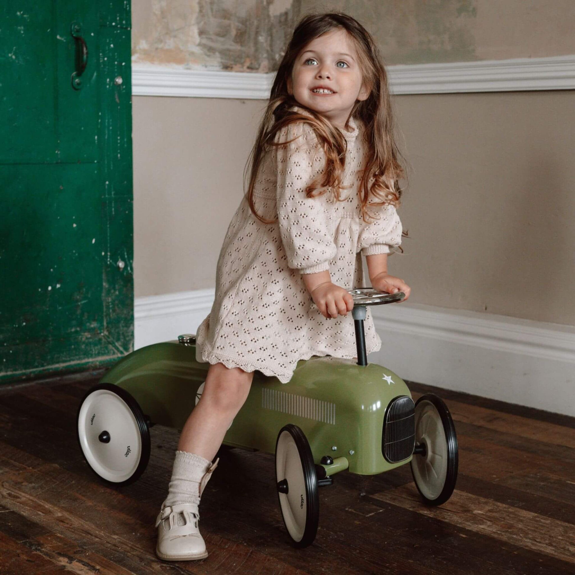 Child riding the Vilac Classic Metal Car in khaki green with a white seat, silver steering wheel, and star emblem, in a rustic interior setting.