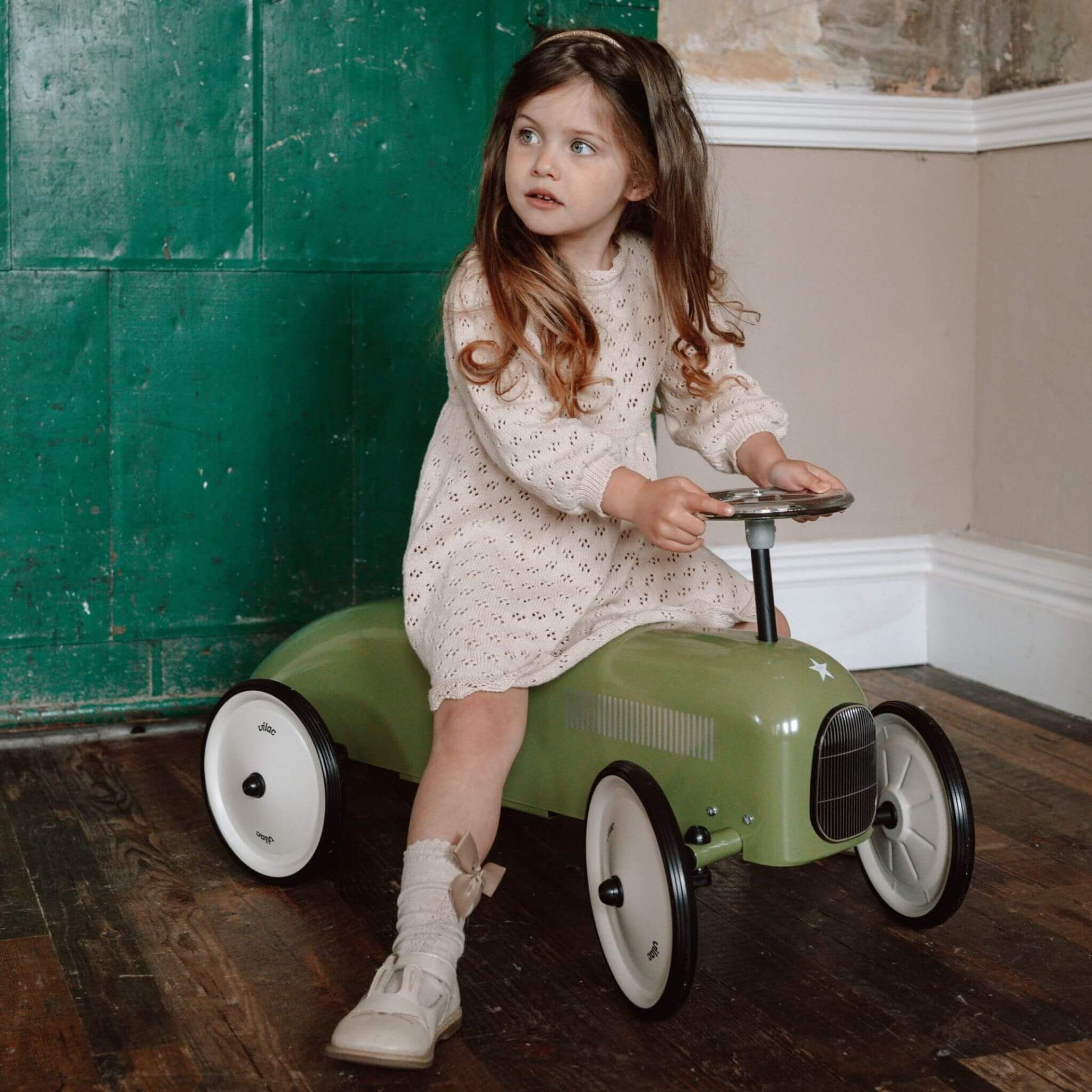 Child sitting on the Vilac Classic Metal Car in khaki green with a white seat, silver steering wheel, and star emblem, in a cosy rustic room.