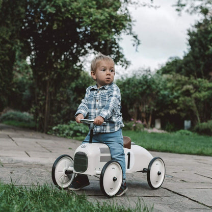 Child riding the Vilac Classic Metal Car in off-white with a brown seat and silver steering wheel, outdoors in a lush garden setting.