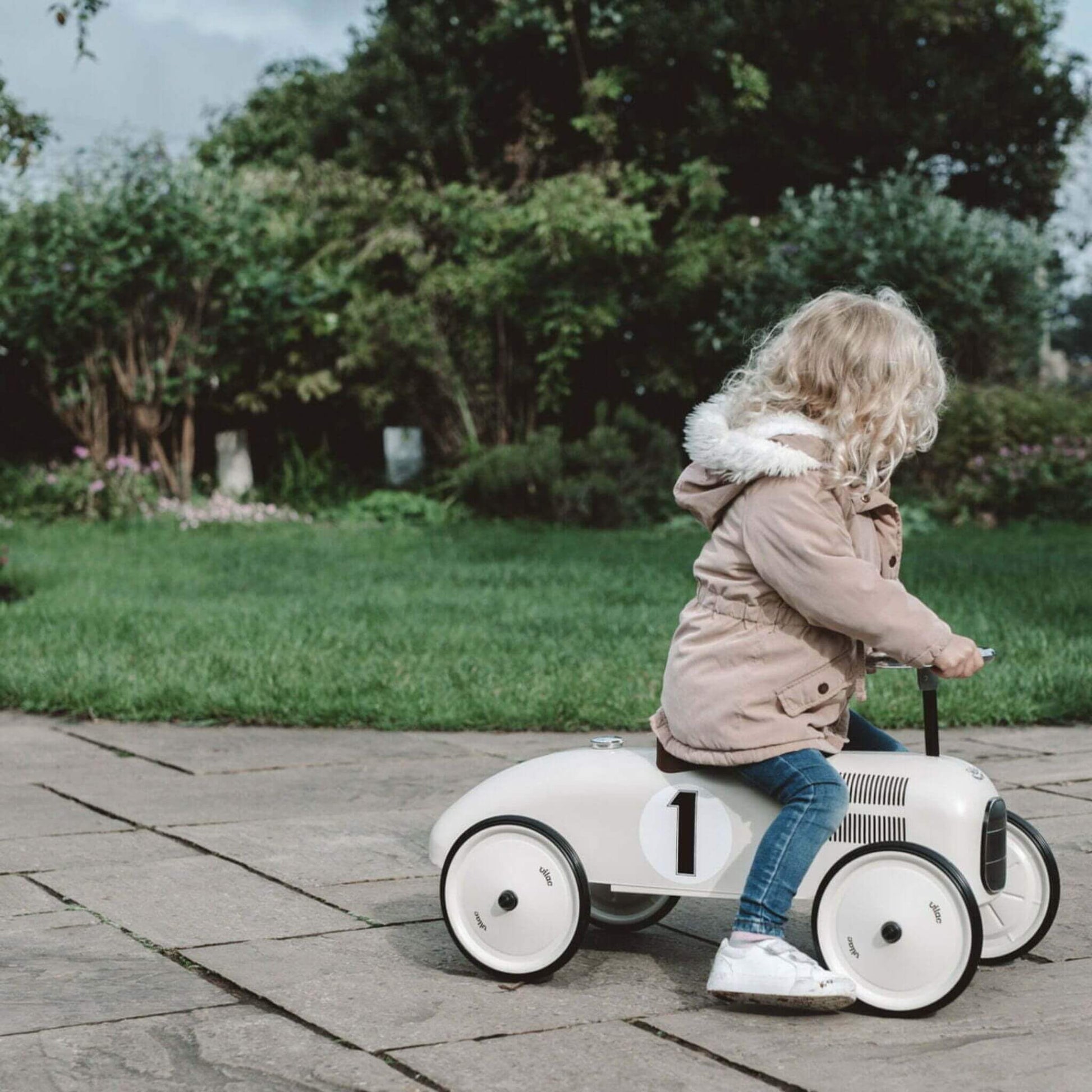 Child riding the Vilac Classic Metal Car in off-white with a brown seat and number 1 detail, outdoors on a garden path surrounded by greenery.