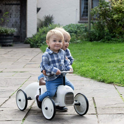 Two children riding the Vilac Classic Metal Car in off-white with a brown seat and silver steering wheel, enjoying playtime outdoors on a garden path.