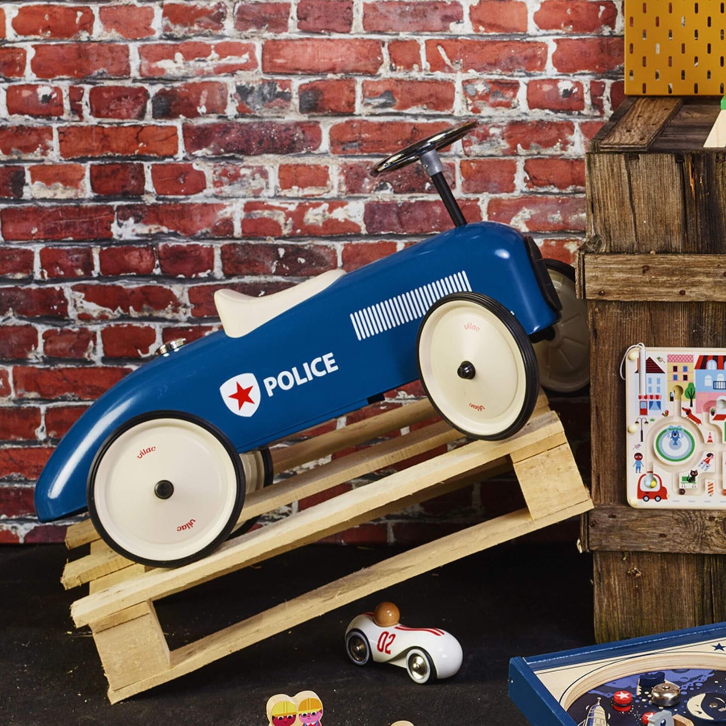 Vilac Classic Metal Car in blue with a white seat and police-themed details, displayed on a wooden ramp against a rustic brick wall, surrounded by toys.