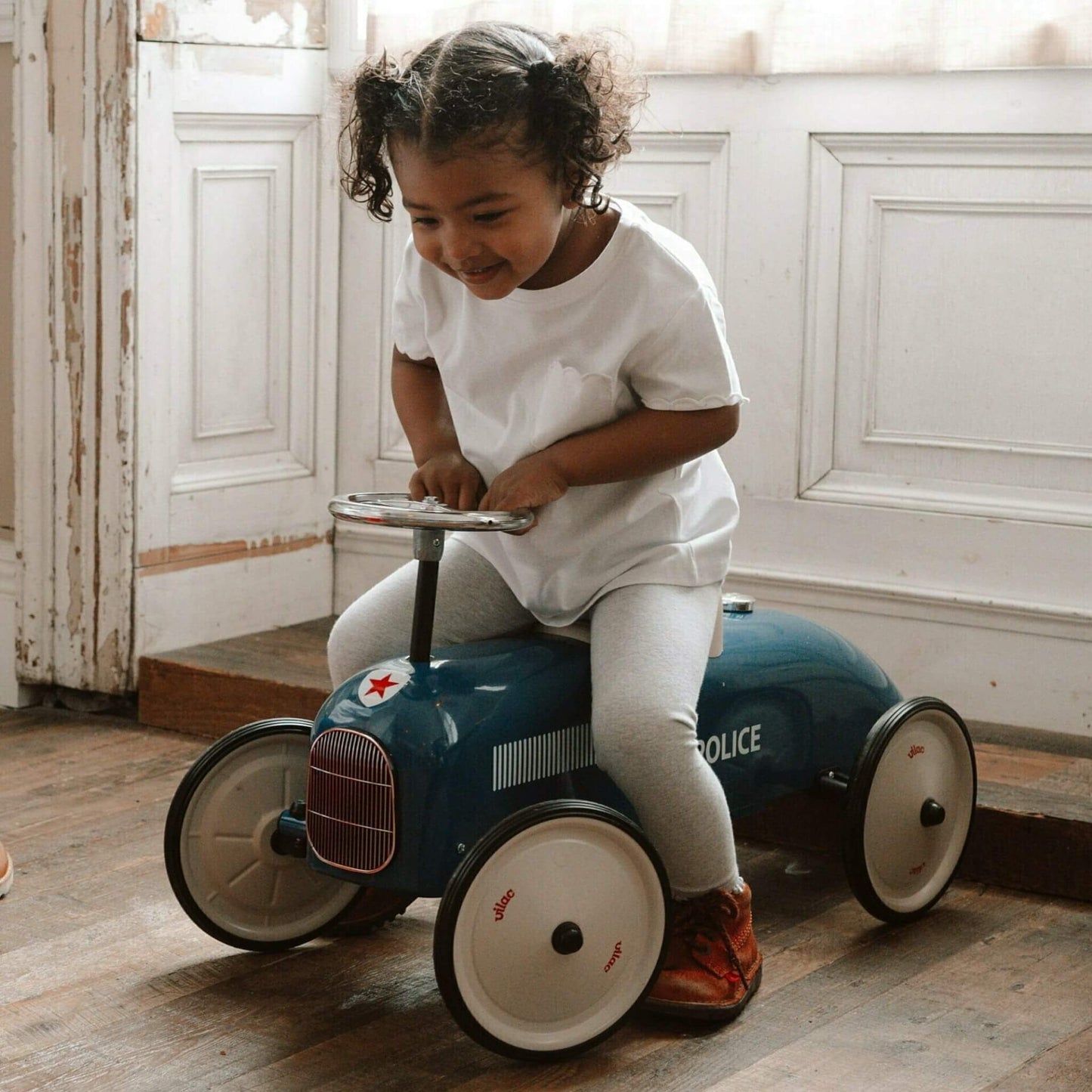 Little girl riding the Vilac Classic Metal Car in blue with a white seat and police-themed design, smiling joyfully in a cosy indoor setting.