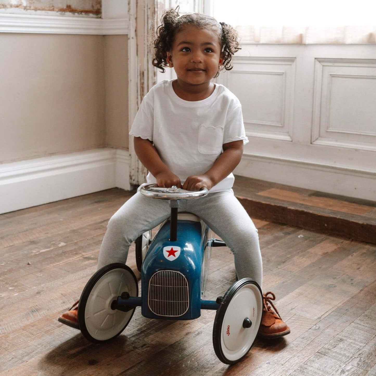 Little girl sitting on the Vilac Classic Metal Car in blue with a white seat and police-themed design, looking content in a cosy indoor space.