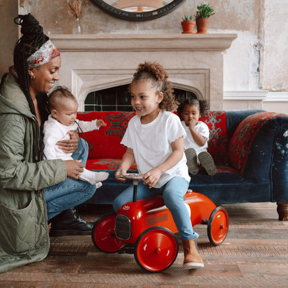 Child riding the Vilac Classic Metal Car in red with a brown seat, surrounded by family in a cosy living room featuring a vintage sofa and decor.