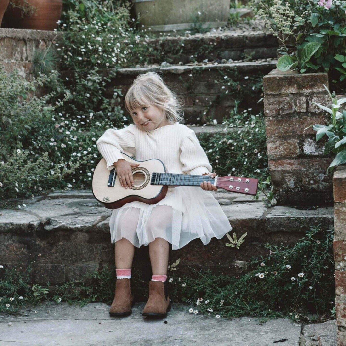 A young girl playing the Vilac natural wood guitar outdoors, seated on stone steps surrounded by greenery.