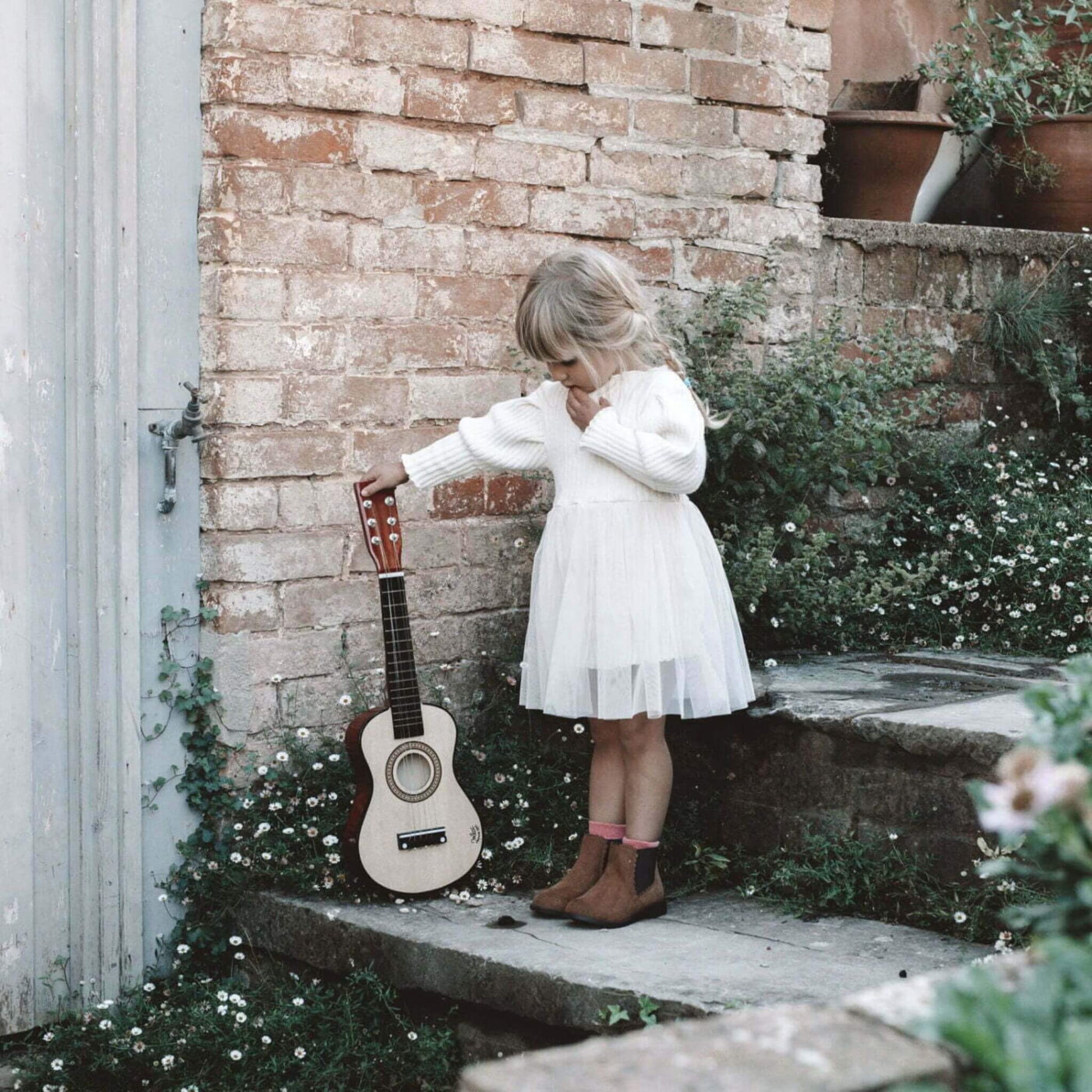 A young girl standing outdoors, gently holding the Vilac natural wood guitar, surrounded by rustic brick walls and greenery. 