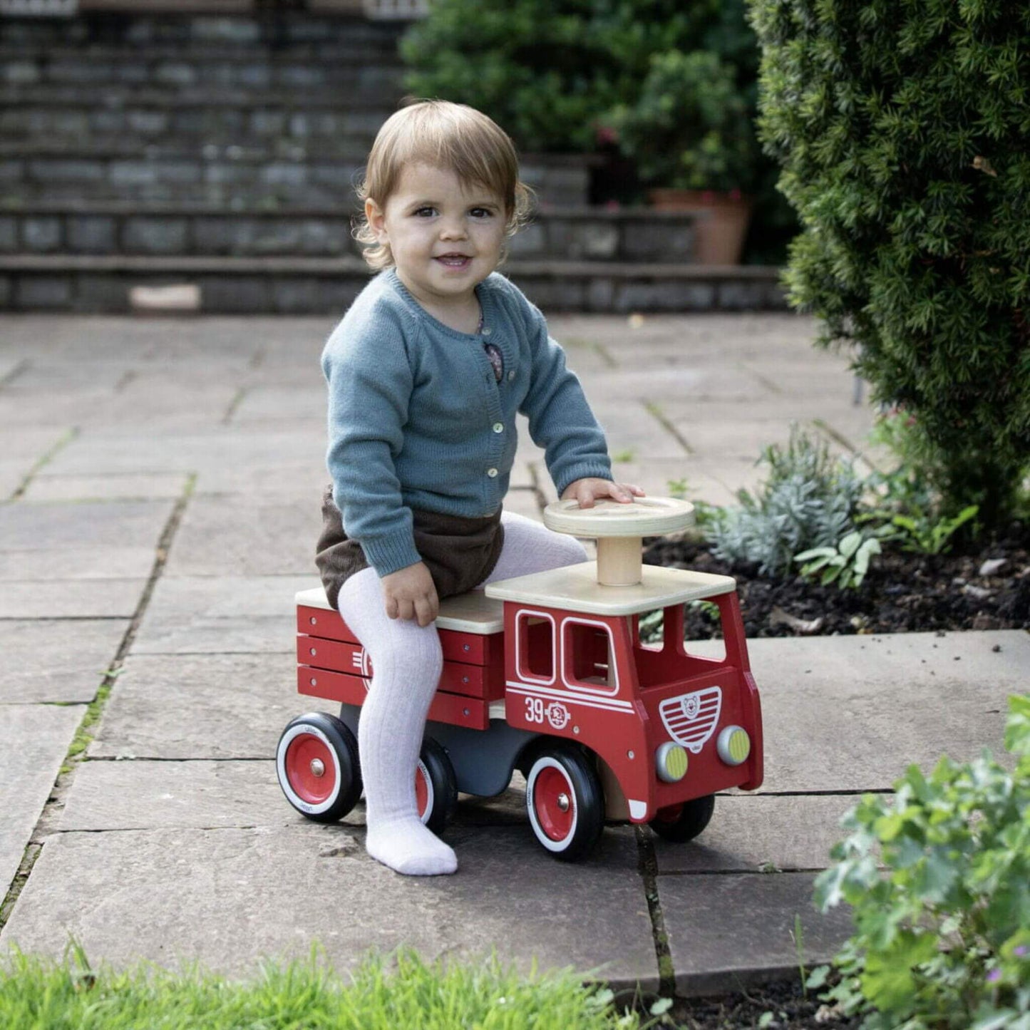 A toddler wearing a blue cardigan, brown shorts, and white tights sits happily on the Vilac Ride-On Fire Truck in a garden setting.