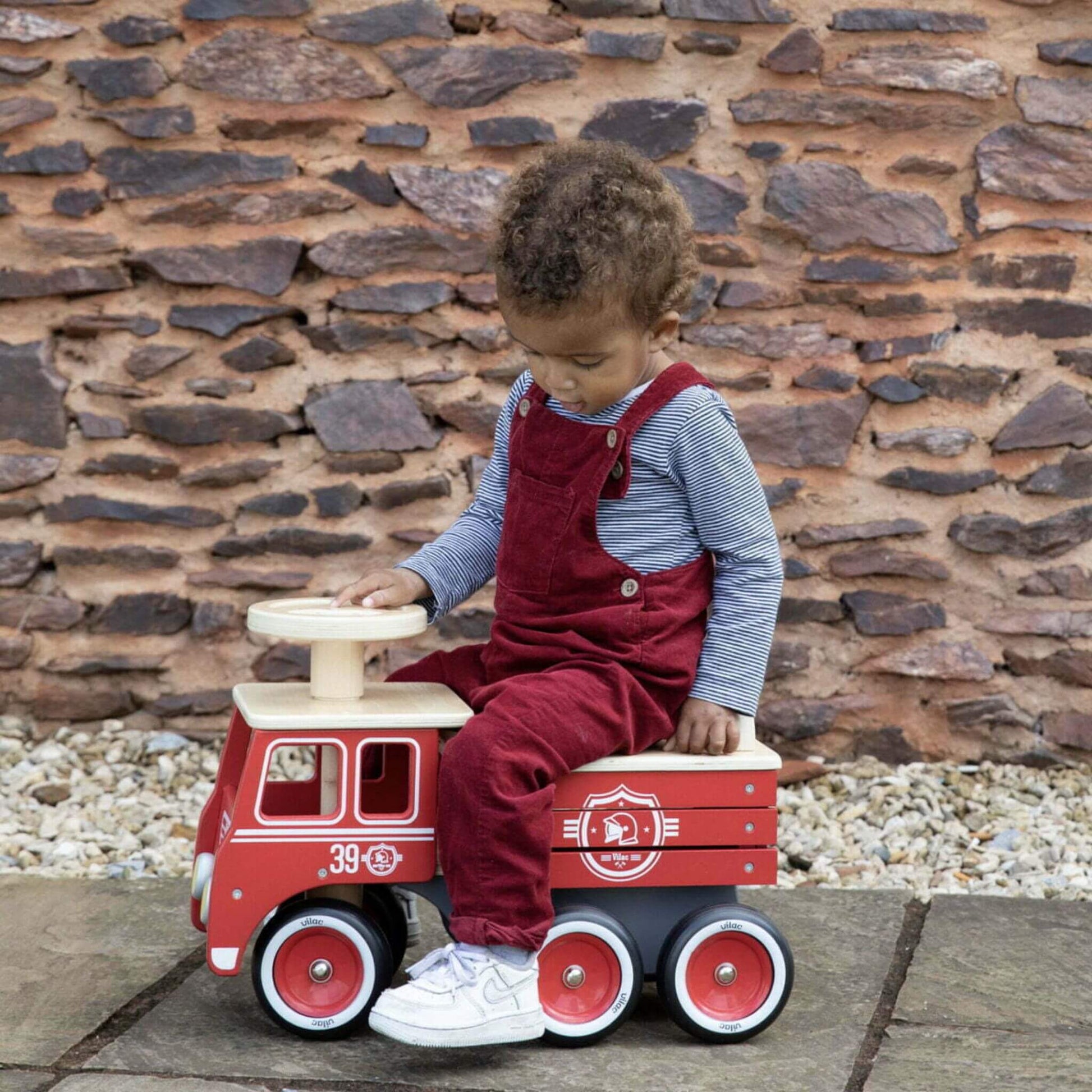 A child dressed in a striped top and red dungarees sits on the Vilac Ride-On Fire Truck, holding the wooden steering wheel, against a rustic stone wall background.