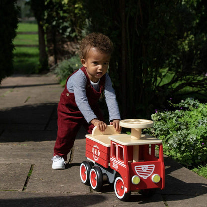 A child wearing red dungarees and a striped top pushes the Vilac Ride-On Fire Truck along a garden path surrounded by greenery and sunlight.