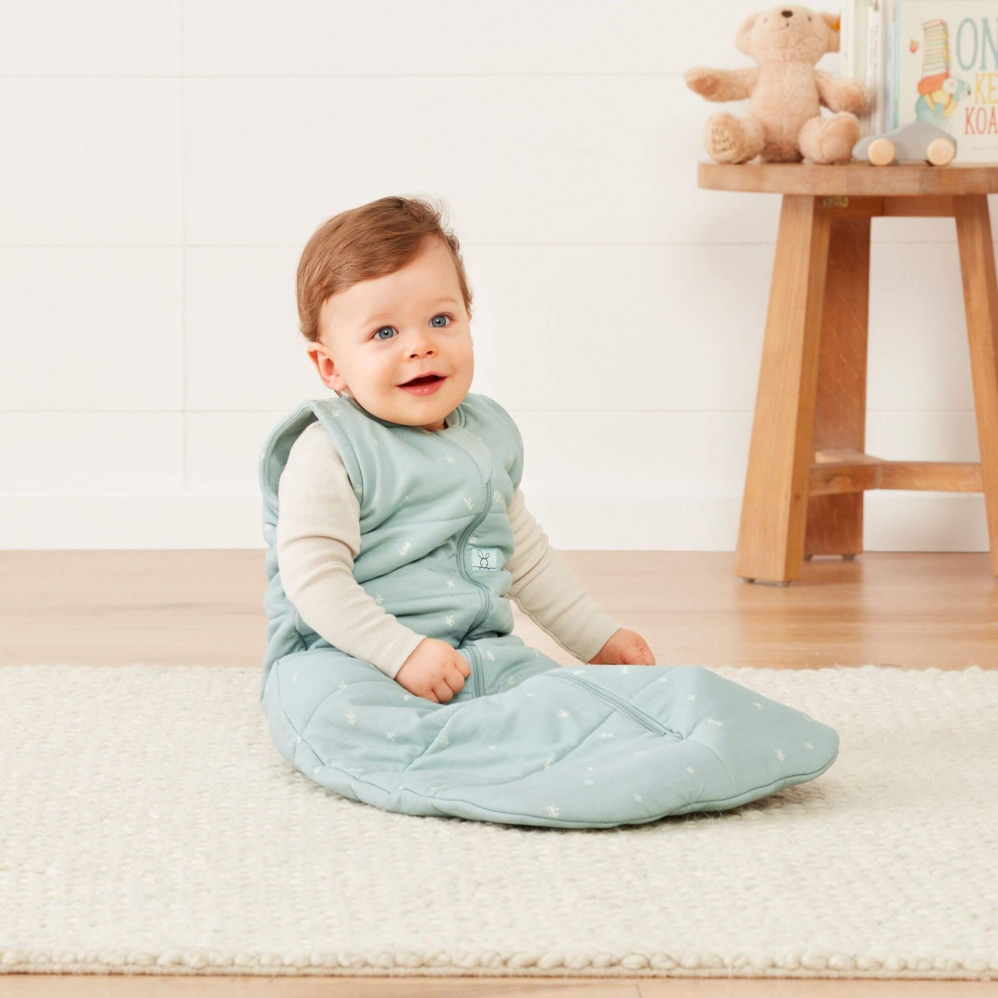 A baby sits on a soft rug wearing a sage green ergoPouch winter jersey sleeping bag, 2.5 TOG, in a cosy nursery with a wooden stool nearby.