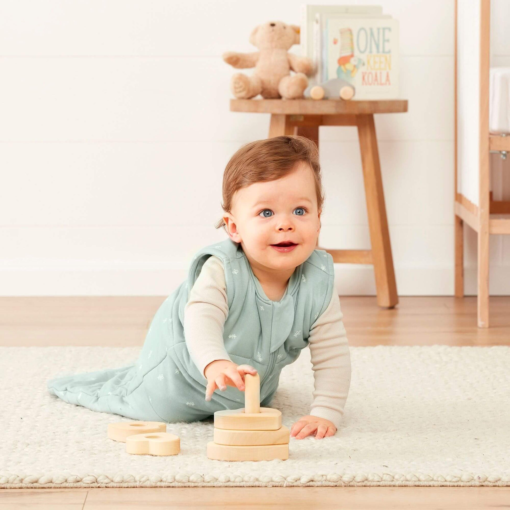 A baby crawls while wearing a sage green ergoPouch winter jersey sleeping bag, 2.5 TOG, playing with wooden stacking toys in a cosy nursery.