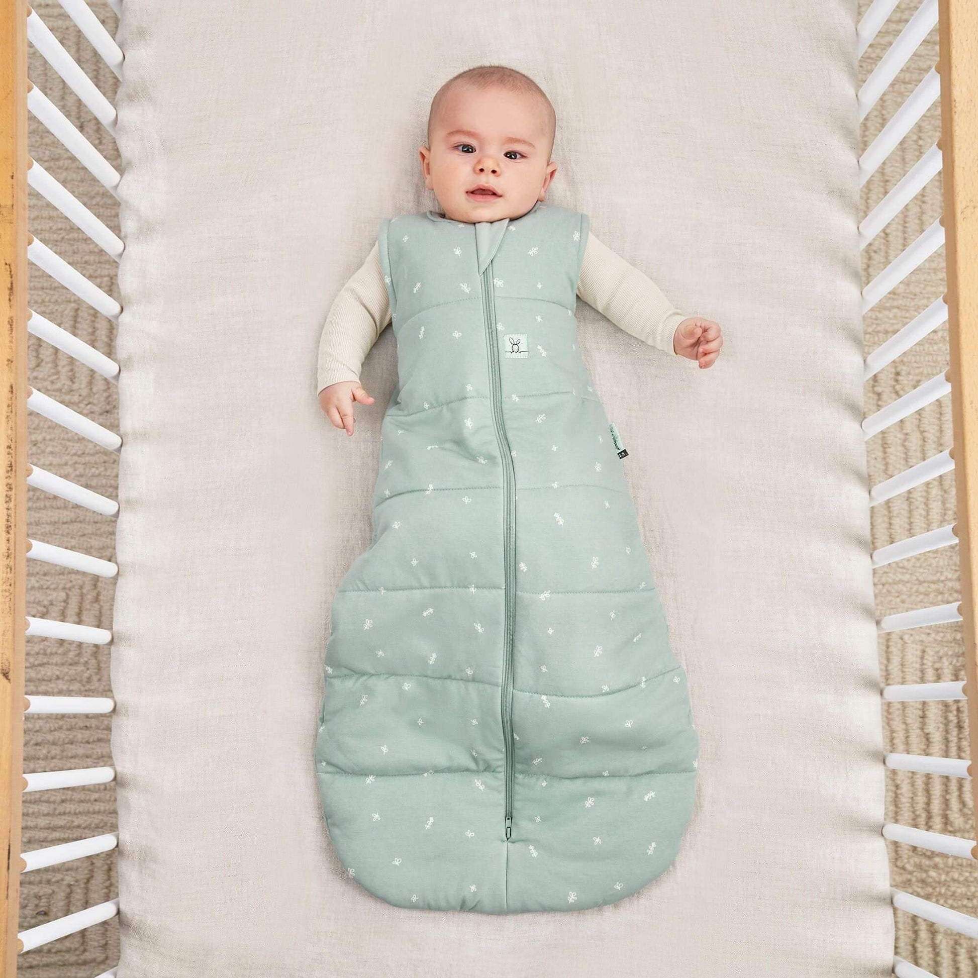 A baby lies in a cot wearing a sage green ergoPouch winter jersey sleeping bag, 2.5 TOG, with a central zip and small white patterns.