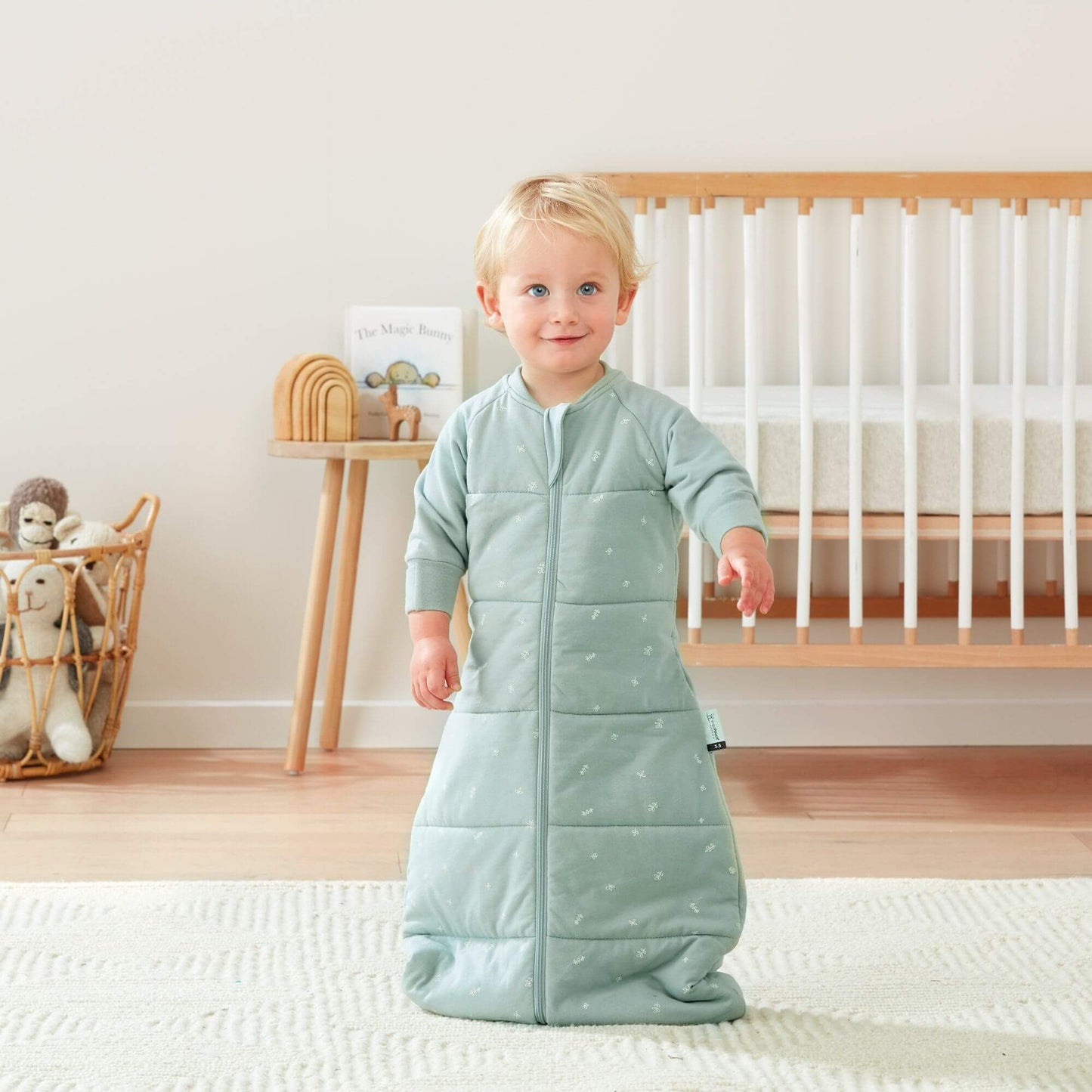  A toddler stands wearing a sage green ergoPouch winter jersey sleeping bag, 3.5 TOG, in a nursery setting with a cot and toys nearby.