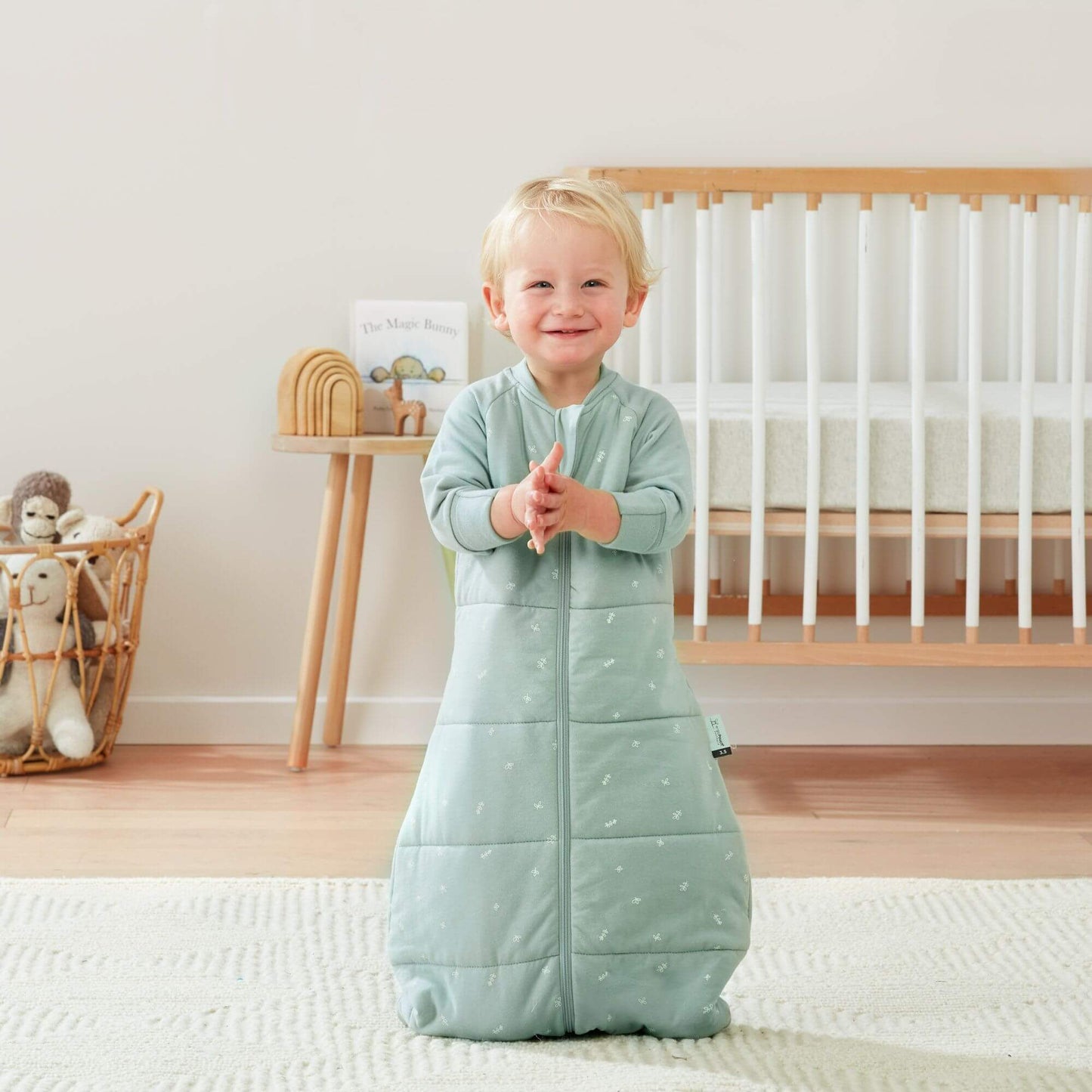 A smiling toddler claps while wearing a sage green ergoPouch winter jersey sleeping bag, 3.5 TOG, in a nursery with a cot and toys.