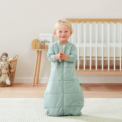 A smiling toddler claps while wearing a sage green ergoPouch winter jersey sleeping bag, 3.5 TOG, in a nursery with a cot and toys.