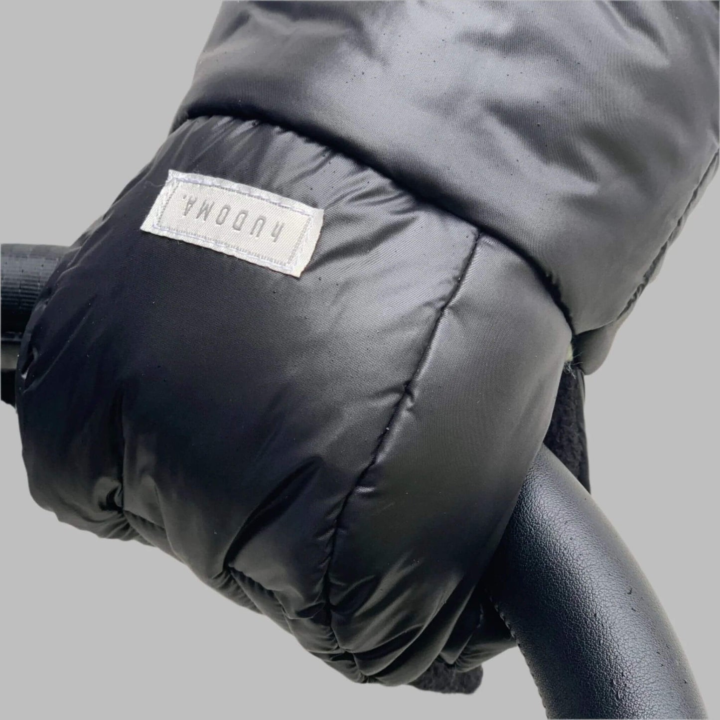 Close-up of the hUDOMA Chelsea Black Pram Glove, highlighting its durable black exterior, soft texture, and branded label, securely wrapped around a stroller handle for cold-weather comfort and convenience.